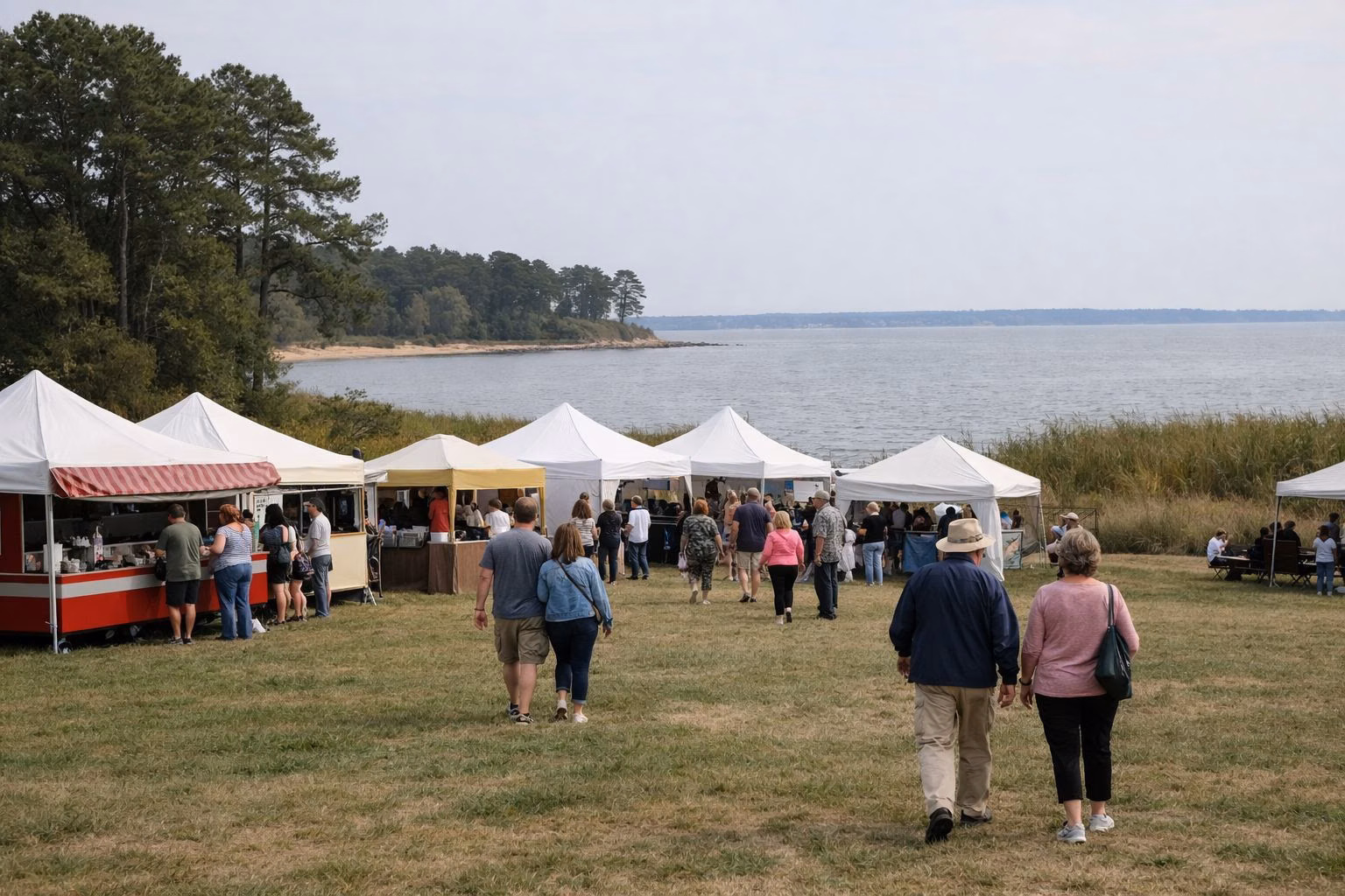 White festival tents and food stands on a large grassy field near the water, people walking between them