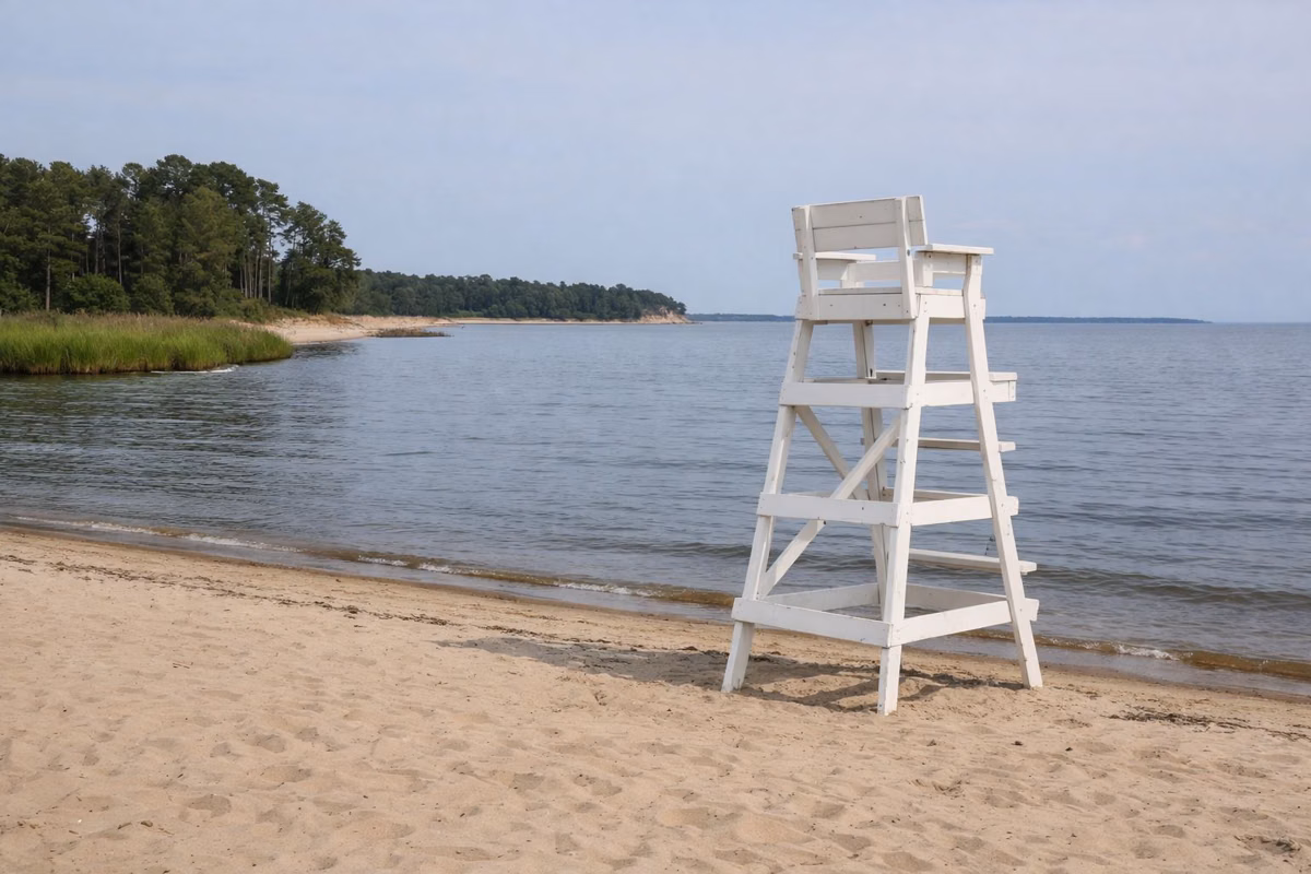 A tall white wooden lifeguard stand on an empty stretch of sand, calm bay water behind it