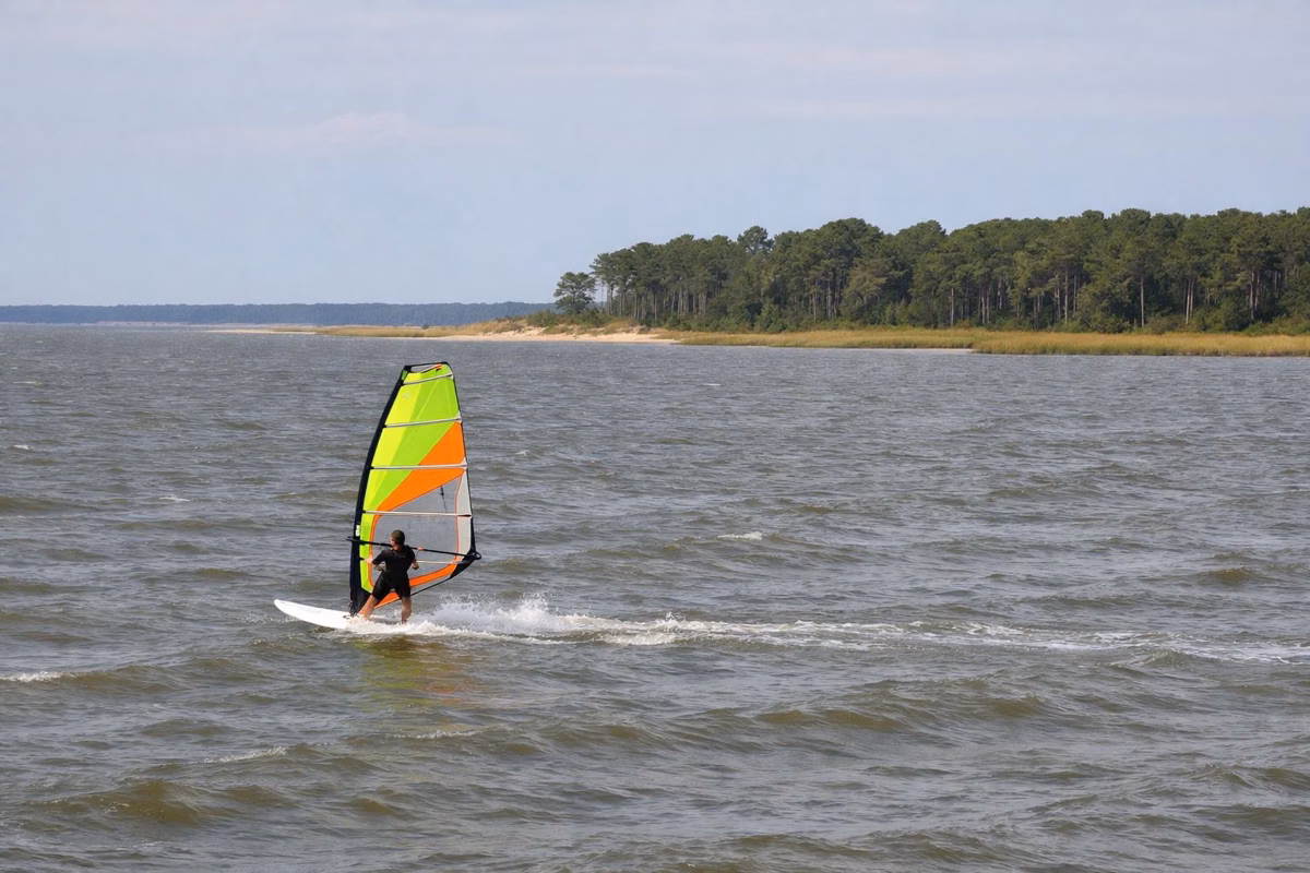 A windsurfer with a bright sail gliding across choppy open bay water, shoreline far behind