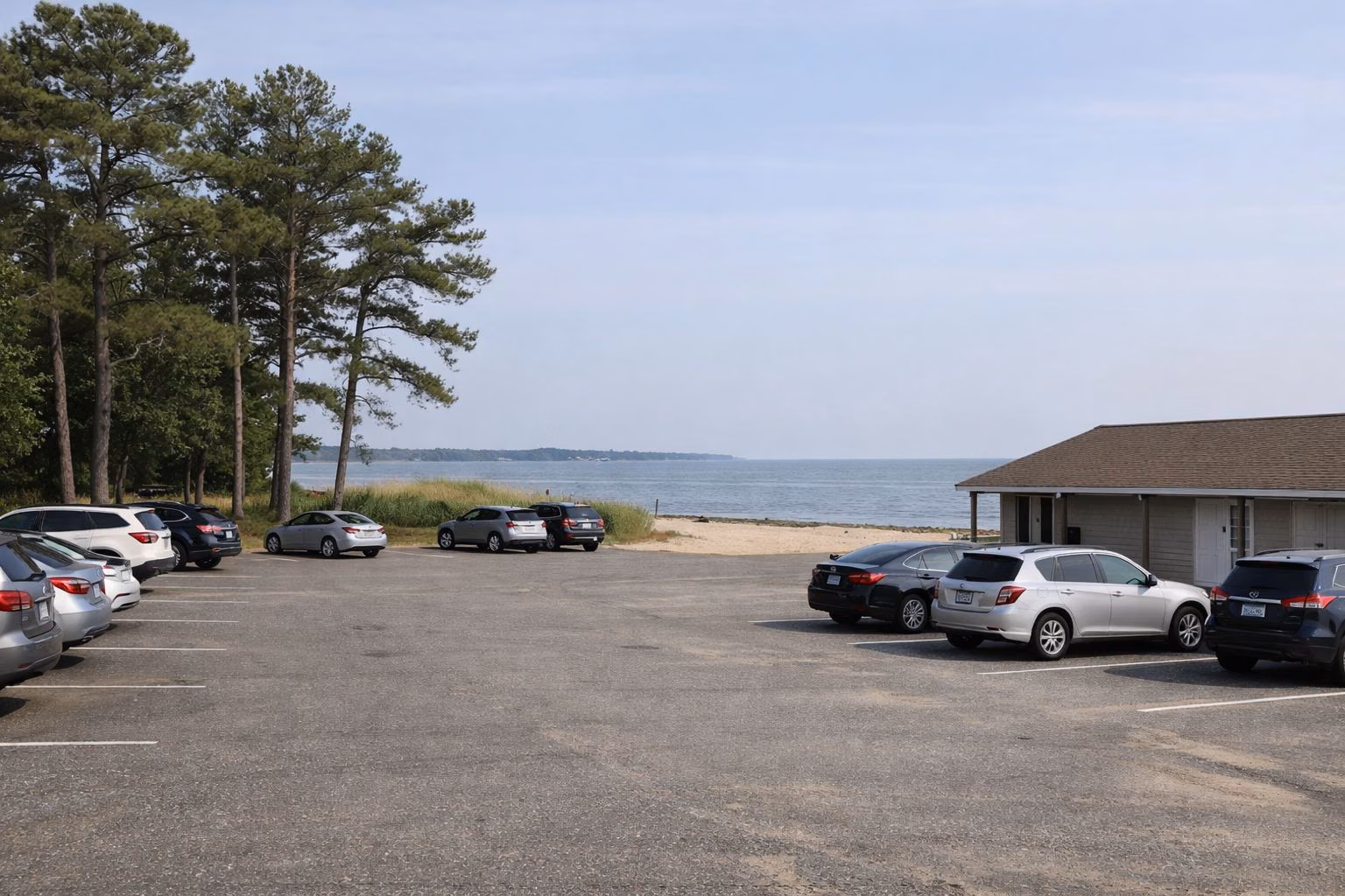 A large paved beach parking lot with parked cars, pine trees and a low bathhouse building at the edge