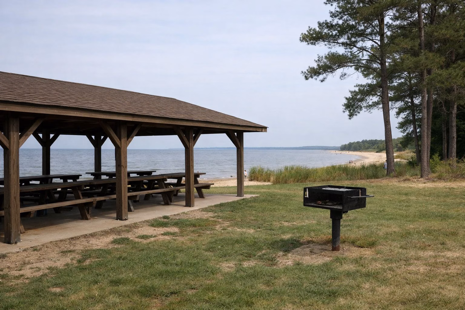 An open-sided wooden picnic pavilion with long picnic tables and a grill beside it on a lawn near a small beach