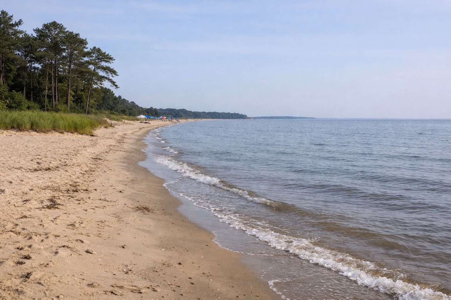 A wide sandy bay beach with gentle small waves, a few beach umbrellas far down the shore, open water to the horizon