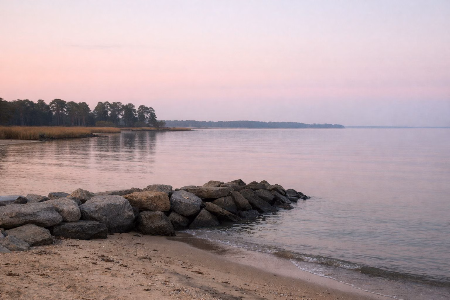 Calm open bay water at dawn seen from a sandy shore with a low rock jetty in the foreground, soft pink sky